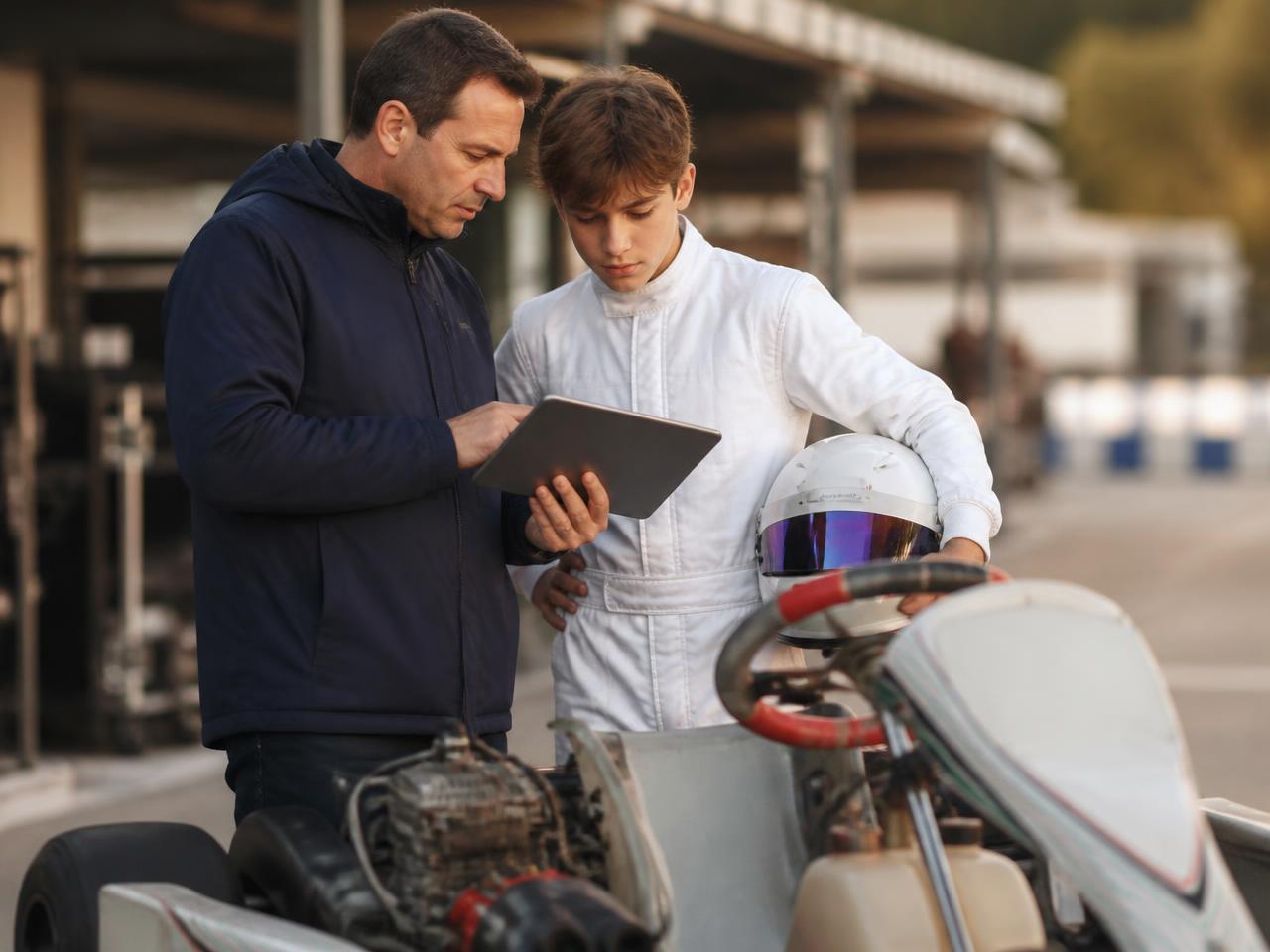 A coach and a young driver in a race suit reviewing data on a tablet beside a race kart